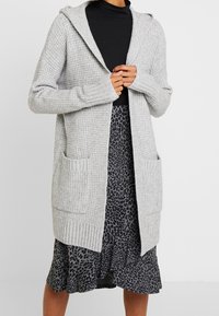 Light grey knit cardigan with a hood, ribbed cuffs, and two front pockets, worn over a patterned black and grey layered dress.