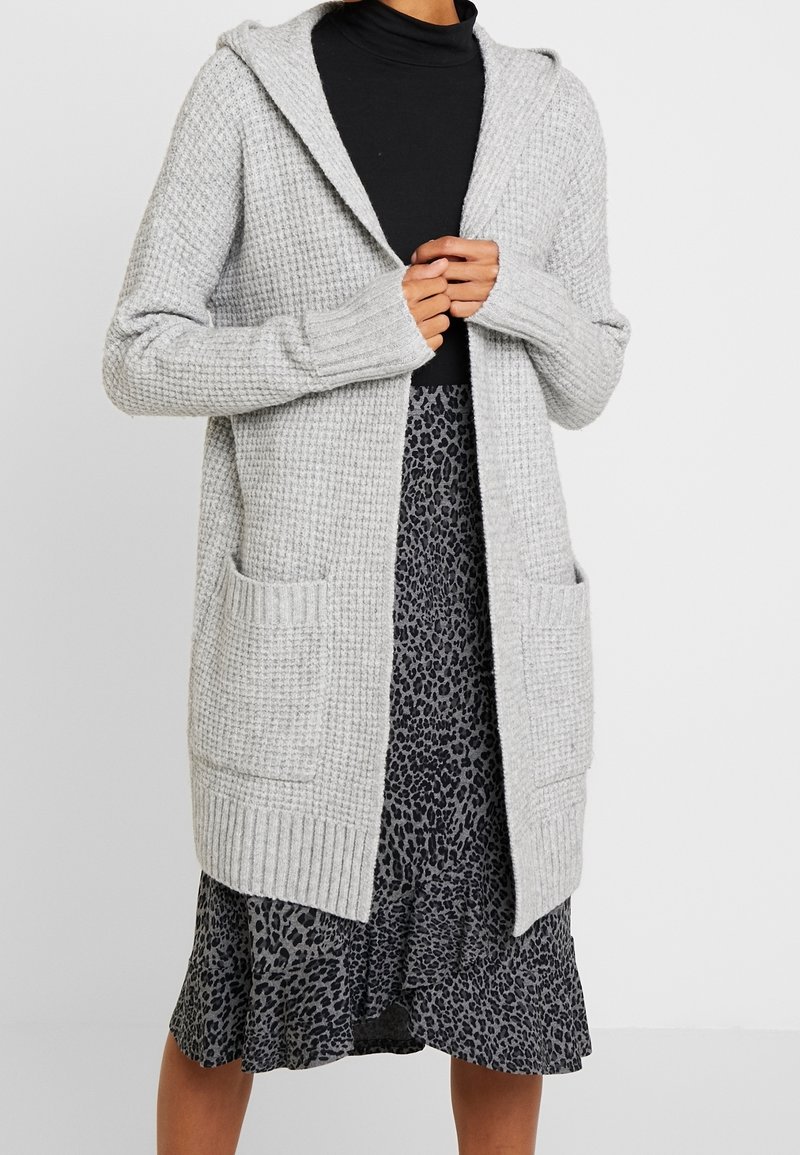Light grey knit cardigan with a hood, ribbed cuffs, and two front pockets, worn over a patterned black and grey layered dress.