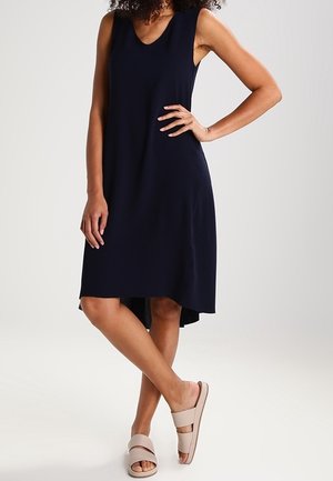 Frau trägt ein ärmelloses, marineblaues knielanges Kleid und beige Pantoletten, steht mit einer Hand in der Hüfte und überschlagenen Beinen.