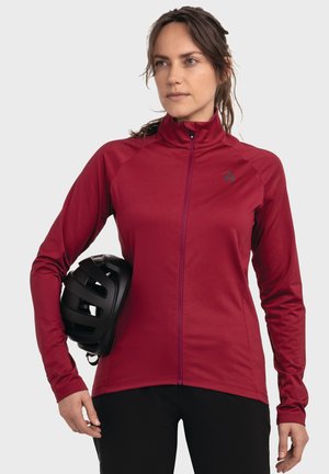 Red, long-sleeve cycling jacket with a high collar, full zipper, and snug fit, paired with a black bicycle helmet held under the arm.