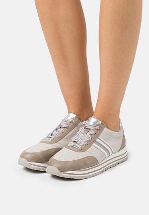 Beige en witte sneakers met wit gestreepte veters, gedragen aan de voeten, met metallic zilveren accenten en gestreepte plateauzolen.