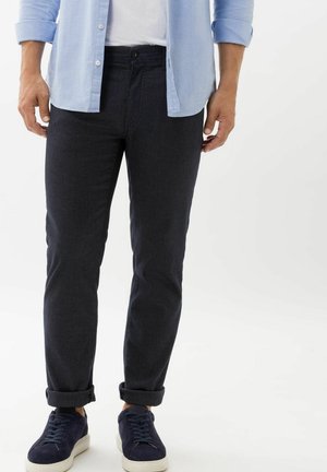 Dark blue, tapered pants with a smooth texture, featuring a button closure, belt loops, and rolled cuffs, paired with navy sneakers.