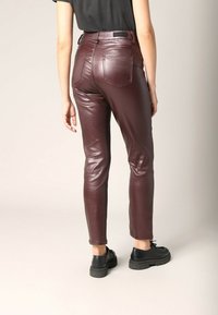Bordeaux faux leren broeken met een slim fit, middelhoge taille en achterzakken. Het model draagt zwarte schoenen, zichtbaar van de enkel.