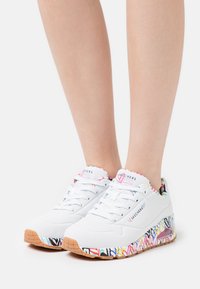 Weiße Sportschuhe mit bunten, gemusterten Akzenten an der Sohle. Obermaterial aus Mesh, runde Zehenpartie und abgerundete Ferse. Mit Schnürsenkeln und gepolstertem Kragen.