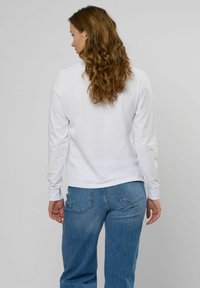 Sieviete ar gariem cirtainiem brūniem matiem, valkājot baltu krekla ar garām piedurknēm un zilas džinsas, stāvot ar muguru pret vienkāršu fonu.