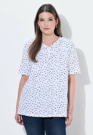 Femme aux longs cheveux bruns portant un chemisier blanc à manches courtes avec de petits motifs de cœurs noirs et un jean bleu foncé, debout contre un mur blanc.