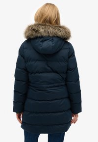 Giacca a vento blu navy con cappuccio foderato in pelliccia, design trapuntato e vita con cintura, con maniche lunghe e lunghezza fino a metà coscia.