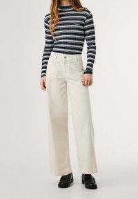 Långärmad topp med marinblå ränder och hög krage, tillsammans med vida, krämfärgade byxor och svarta skor, allt på en neutral bakgrund.