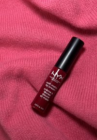 NYX Soft Matte Lip Cream i ett rött rör med ett matt svart lock. Positionerad på en texturerad rosa stickad tygbakgrund.