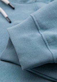 Lys blå sweatshirtærme med ribbet manchet og hvide snore på blød stoffundgrund, der viser tekstur og syningsdetaljer.