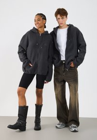 Dva modelky mají na sobě tmavě šedé fleece mikiny se zipem. Jeden modelčka kombinovala s černými cyklistickými šortkami a vysokými černými botami, druhá s vybledlými volnými džíny a teniskami.