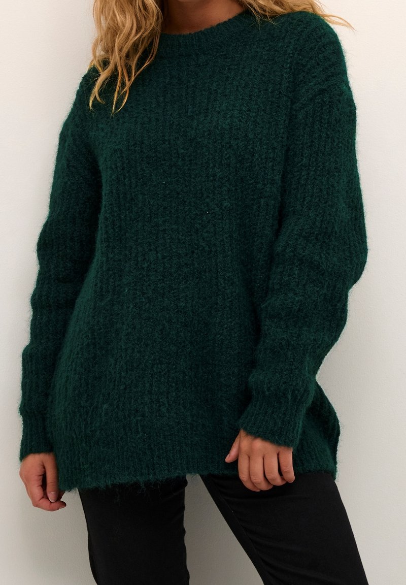 Vrouw in een donkergroene, fluffy gebreide trui met lange mouwen, gecombineerd met zwarte broek, staand tegen een egale lichte muur.
