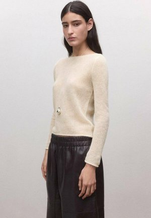 Joven con cabello largo y oscuro que lleva un suéter de punto beige, pantalones de cuero negros con cintura elástica y un collar largo con colgante.