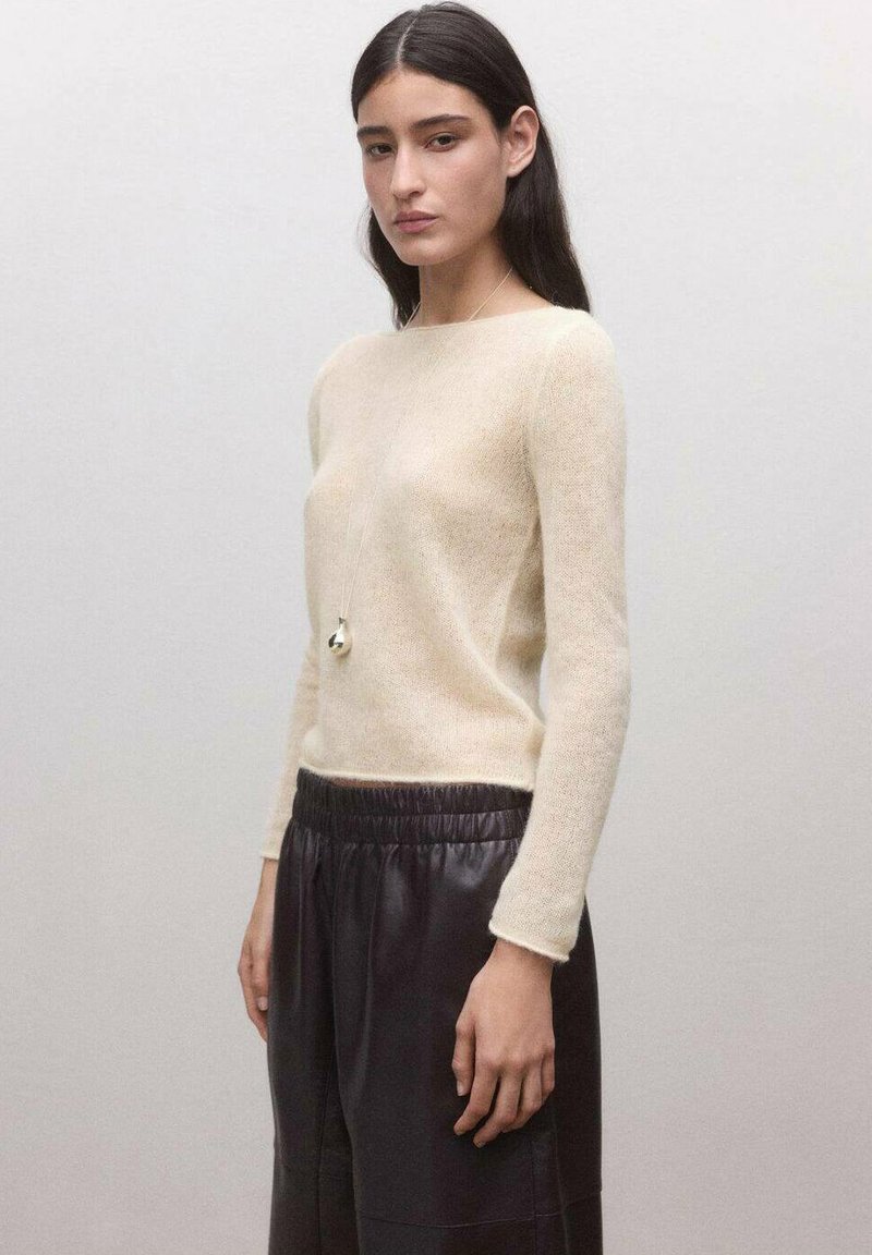 Mlada žena s dugom tamnom kosom nosi bež pleteni džemper, crne kožne hlače s elastičnim pojasom i dugi ogrlicu s privjeskom.