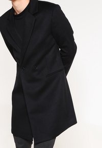 Cappotto in lana nero doppiopetto con una vestibilità su misura, dotato di revers a scallop, tasche laterali e tessuto leggermente strutturato.