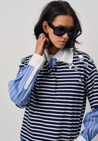 Gestreepte polo t-shirt met marineblauwe en witte lijnen, lichtblauwe mouwen, een witte kraag en versieringen op de schouders. Model draagt een donkere zonnebril.