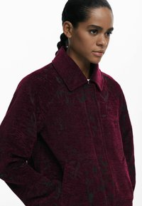 Cappotto bordeaux in tessuto, con colletto e un sottile motivo floreale nero. Vestibilità ampia con tasche laterali e una fodera interna liscia.
