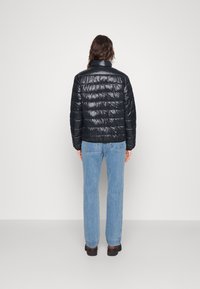 Black puffer jacket with a shiny finish, featuring a stand collar and horizontal quilted pattern, worn with light blue jeans.