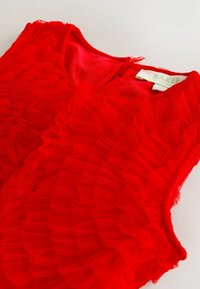 Červený top bez rukávov s vrstvenými volánmi z priesvitnej látky. Výstrih je mierne zaoblený a vzadu je viditeľná visačka.