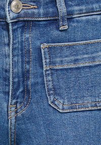 Približnji posnetek modrih jeansa z gumbom na pasu, zanko za pas, sprednjim žepom in podrobnim šivom.