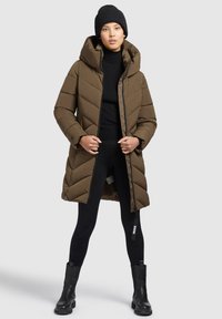 Rjav puffer plašč s kapuco, ki ima diagonalno teksturo in dolg kroj. Obučen čez črno turtleneck majico in legice, v kombinaciji s črnimi čevlji.