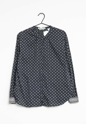 Blouse noire à manches longues avec petits motifs géométriques blancs et poignets contrastants, suspendue sur un cintre noir sur fond blanc.