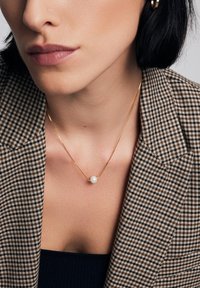 Złoty naszyjnik z małym perełkowym centrałem, noszony na ciemnym topie, w zestawieniu z marynarką w jodełkę, podkreślającą gładką teksturę.