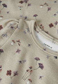 Beiger Stoff mit kleinen lila und braunen Blumen- und Pilzdrucken, mit einem Rundhalsausschnitt und gerafften Details nahe der Schulter.