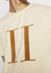 Persona vestida con una camiseta color crema con dos grandes líneas verticales marrones impresas en el pecho, recortada en el cuello y los hombros.