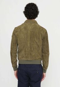 Giacca in suede verde oliva con fascia a coste in vita, dotata di colletto e dettagli sul pannello posteriore. La vista posteriore mostra una finitura testurizzata e un design snello.