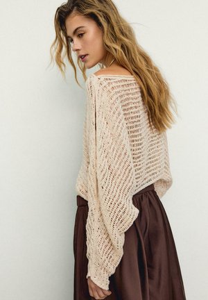 Žena s dlouhými vlnitými vlasy, oblečená v volném béžovém pleteném svetru a tmavě hnědé sukní s vysokým pasem, stojící před jednobarevnou světle zbarvenou zdí.