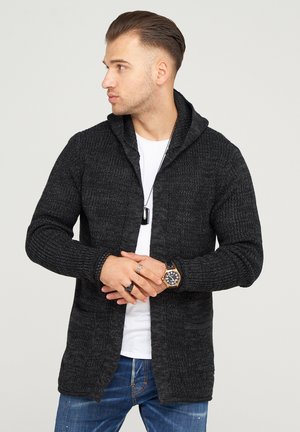 Black knitted cardigan with a shawl collar, open front, two front pockets, and a textured pattern. Worn over a white t-shirt.
