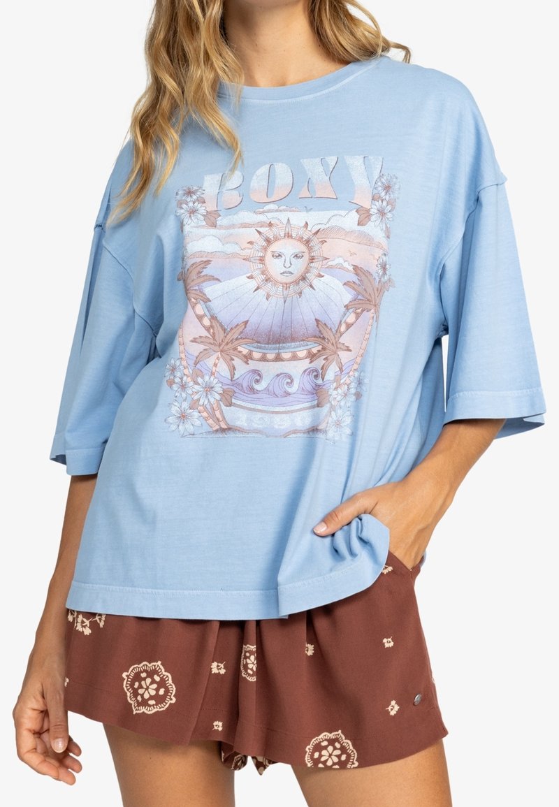 Camiseta oversized de color azul claro con un gráfico de sol y detalles florales, hecha de algodón suave. Combinada con pantalones cortos marrones con estampado.