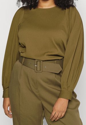 Frau trägt eine olivgrüne Bluse mit langen, aufgepufften Ärmeln und passende, hoch taillierte Hose mit breitem Gürtel.