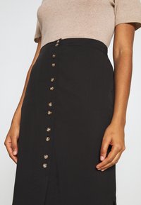 Černá sukně s rovnými liniemi vyrobená z hladkého materiálu. Obsahuje řadu zlatých knoflíků na přední straně a boční rozparek.