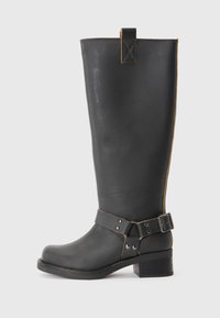 Bota de cuero negro hasta la rodilla con un acabado texturizado, que presenta un acento de hebilla lateral y un tacón bajo. Costura detallada a lo largo de las costuras.