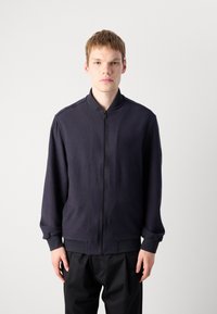 Jonge man met kort haar, gekleed in een navy bomberjack met rits en zwarte broek, staand tegen een effen lichtgrijze achtergrond.