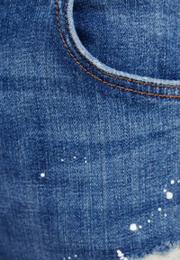 Blå denim med en slitningseffekt, som har fransade kanter och detaljer med vit färgstänk. Orange sömnad runt fickan.