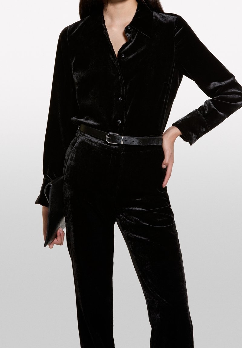 Chemise en velours noir avec col pointu, boutonnée sur le devant, associée à un pantalon taille haute en velours assorti et une ceinture noire.