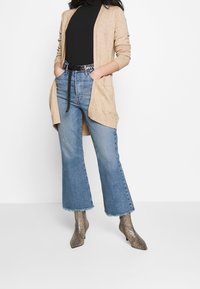 Beige stickad cardigan med öppen front, kombinerad med ljusblå utsvängda jeans och bruna ankelboots i ormskinn. Har en svart bälte vid midjan.