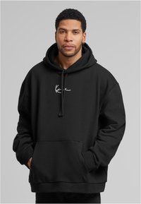 Bărbat cu păr creț scurt, purtând cercei din aur în formă de inel și un hanorac negru cu buzunare frontale și un mic logo alb, cu mâinile în buzunare.