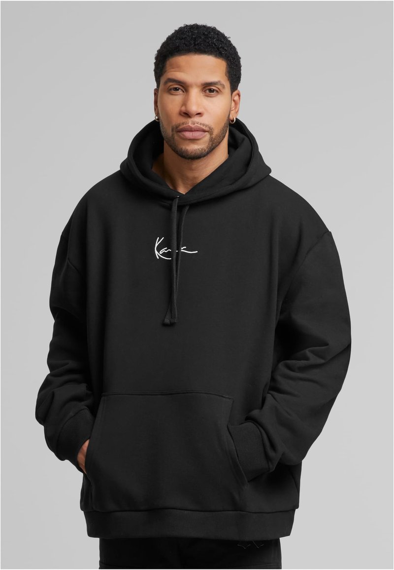 Bărbat cu păr creț scurt, purtând cercei din aur în formă de inel și un hanorac negru cu buzunare frontale și un mic logo alb, cu mâinile în buzunare.