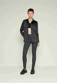 Joven de pie, vistiendo un top corto negro, una camisa negra brillante con botones, jeans skinny oscuros y zapatos de plataforma negros, frente a un fondo liso.