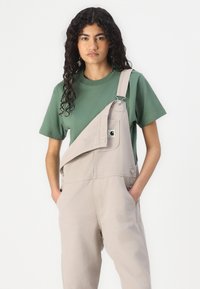 Lichtbeige salopette met één losgeknoopte schouderband, gedragen over een donkergroen T-shirt. Bevat een voorkantzak en is gemaakt van zacht katoen.
