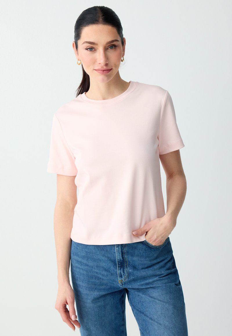 Femme aux cheveux foncés attachés en queue de cheval, portant des boucles d'oreilles créoles dorées, un t-shirt rose clair à manches courtes et un jean bleu, debout avec une main dans la poche.