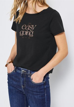 Vrouw draagt een zwart T-shirt met de tekst "The easy going" en blauwe jeans, handen in de zakken en een bruine armband aan de linkerpols.