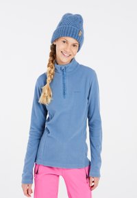 Pullover in pile azzurro chiaro con colletto alto, zip frontale e accenti blu; abbinato a pantaloni rosa brillante. Berretto lavorato a maglia blu in testa.