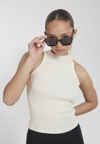 Top de tirantes acanalado en beige claro con un cuello alto, combinado con gafas de sol rectangulares oscuras. Textura suave, forma ajustada y diseño minimalista.