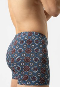 Gros plan sur une personne portant un boxer bleu avec un motif floral circulaire rouge, blanc et marron sur un fond clair.