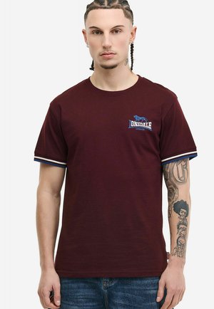 Mężczyzna z zaplecionymi włosami, ubrany w bordową koszulkę Lonsdale z paskowanymi krawędziami rękawów i widocznym tatuażem na prawym ramieniu.